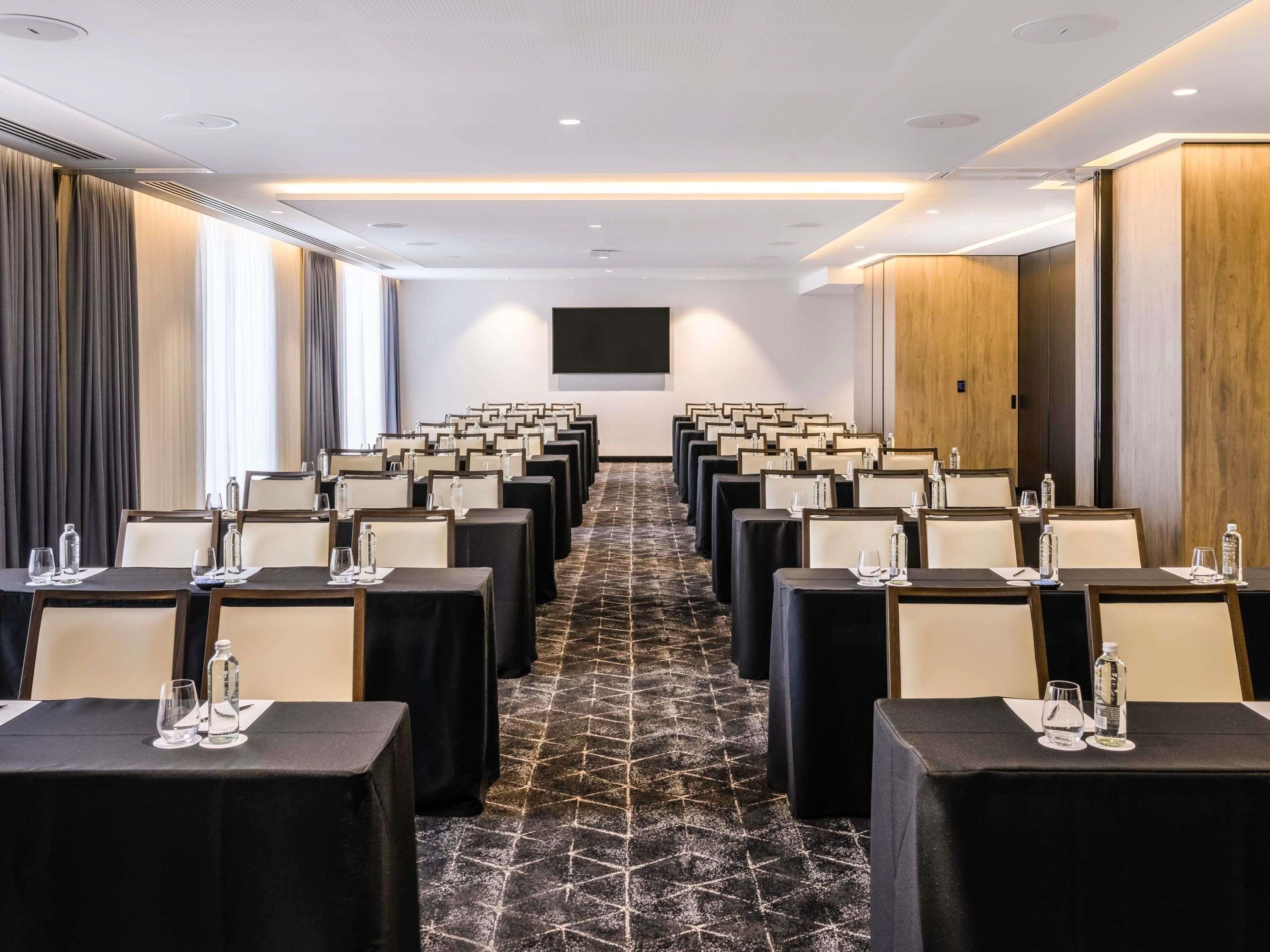 A conference room with rows of tables, chairs, and a large screen at the front.