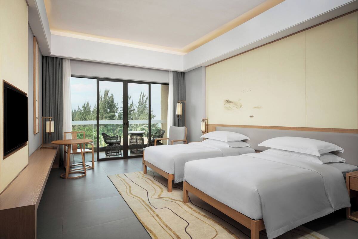 Ein modernes Hotelzimmer mit zwei Betten, einem Schreibtisch, einem Sessel, einem Fernseher und einem Balkon mit Blick auf Bäume.
