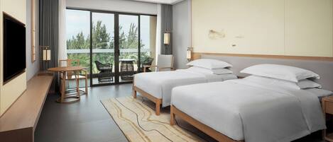 A modern hotel room with two beds, a desk, a chair, a TV, and a balcony with a view of trees.