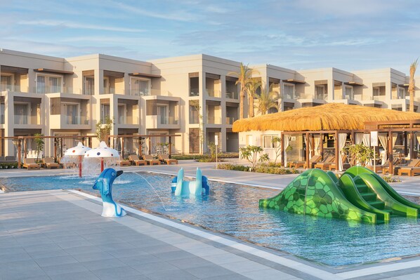 A swimming pool with dolphin sculptures and slides, surrounded by modern buildings with balconies and lounge chairs.