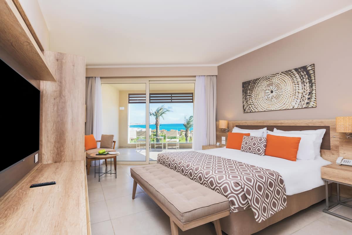 Quarto de hotel com cama, um banco, televisão, escrivaninha e vista para o oceano.