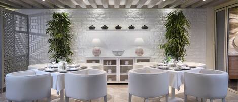 A dining area with white tables and chairs, a stone wall, and potted plants.