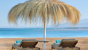 Une plage avec deux chaises longues en bois, placées sous un parasol en chaume, d’où l’on peut admirer une mer calme et des montagnes lointaines.
