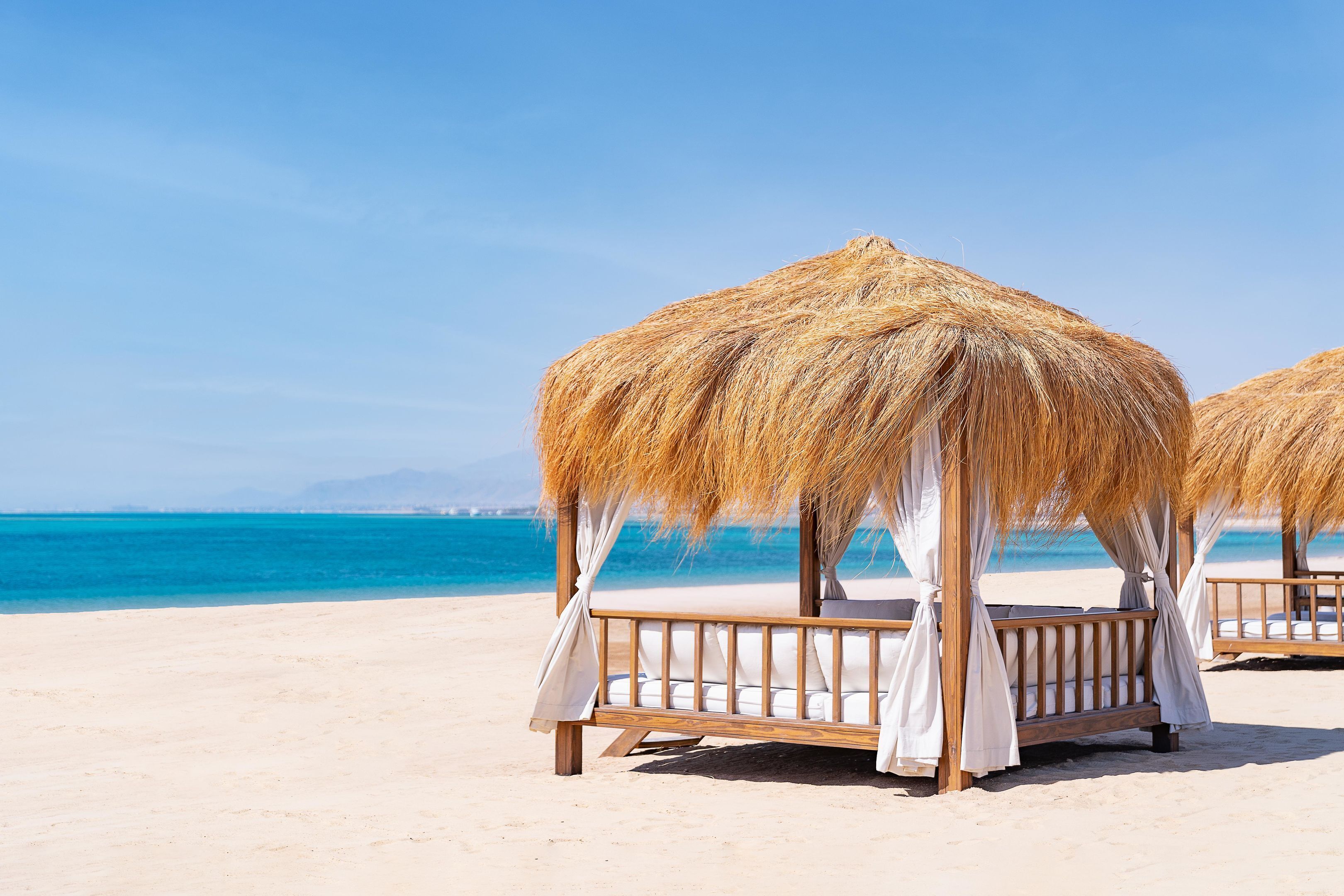 Two thatched beach huts with white curtains, wooden railings, and white cushions on a sandy beach with a clear blue sea and distant mountains.