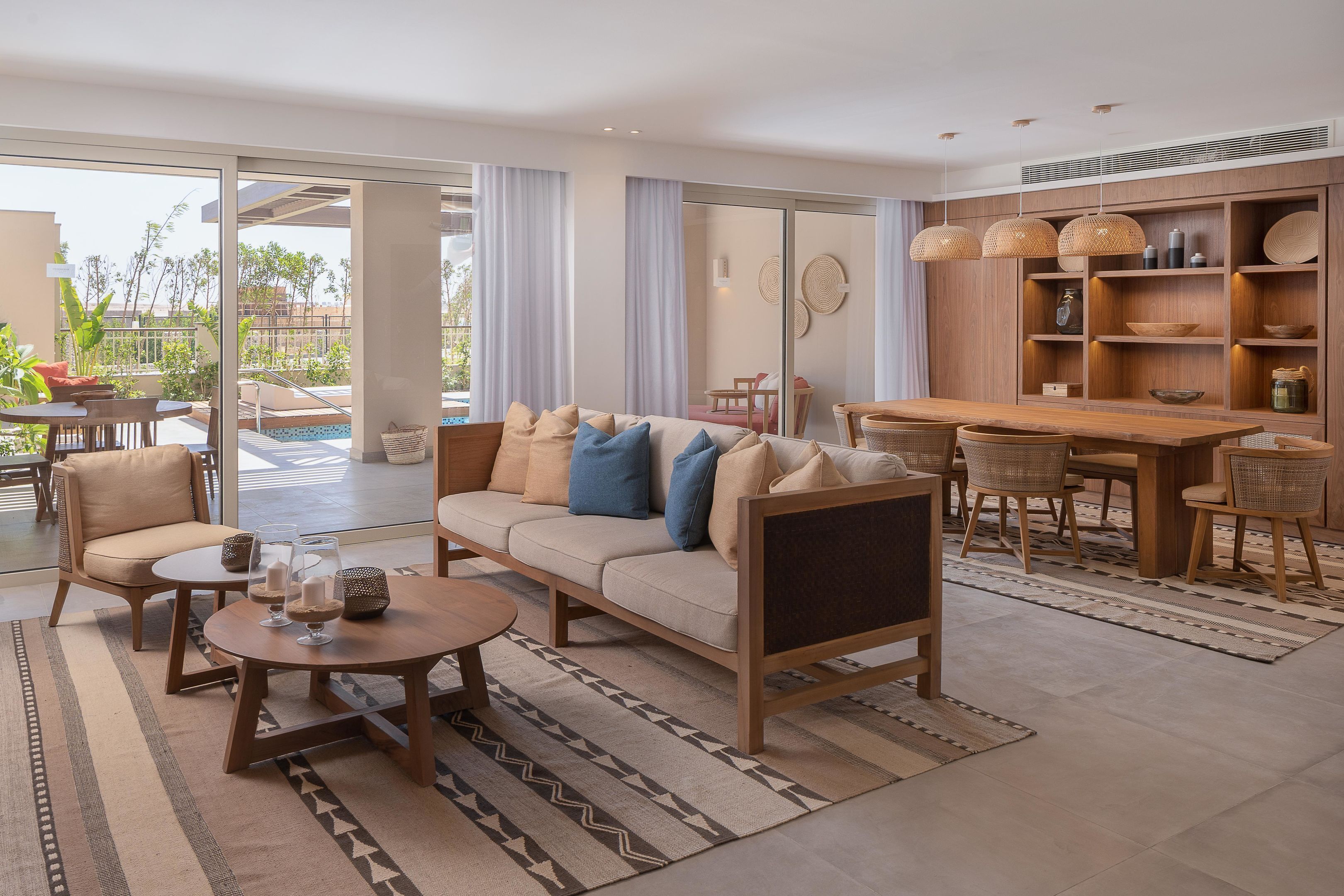 A modern living room with a sofa, coffee table, and dining area, featuring a large sliding glass door to an outdoor pool area.