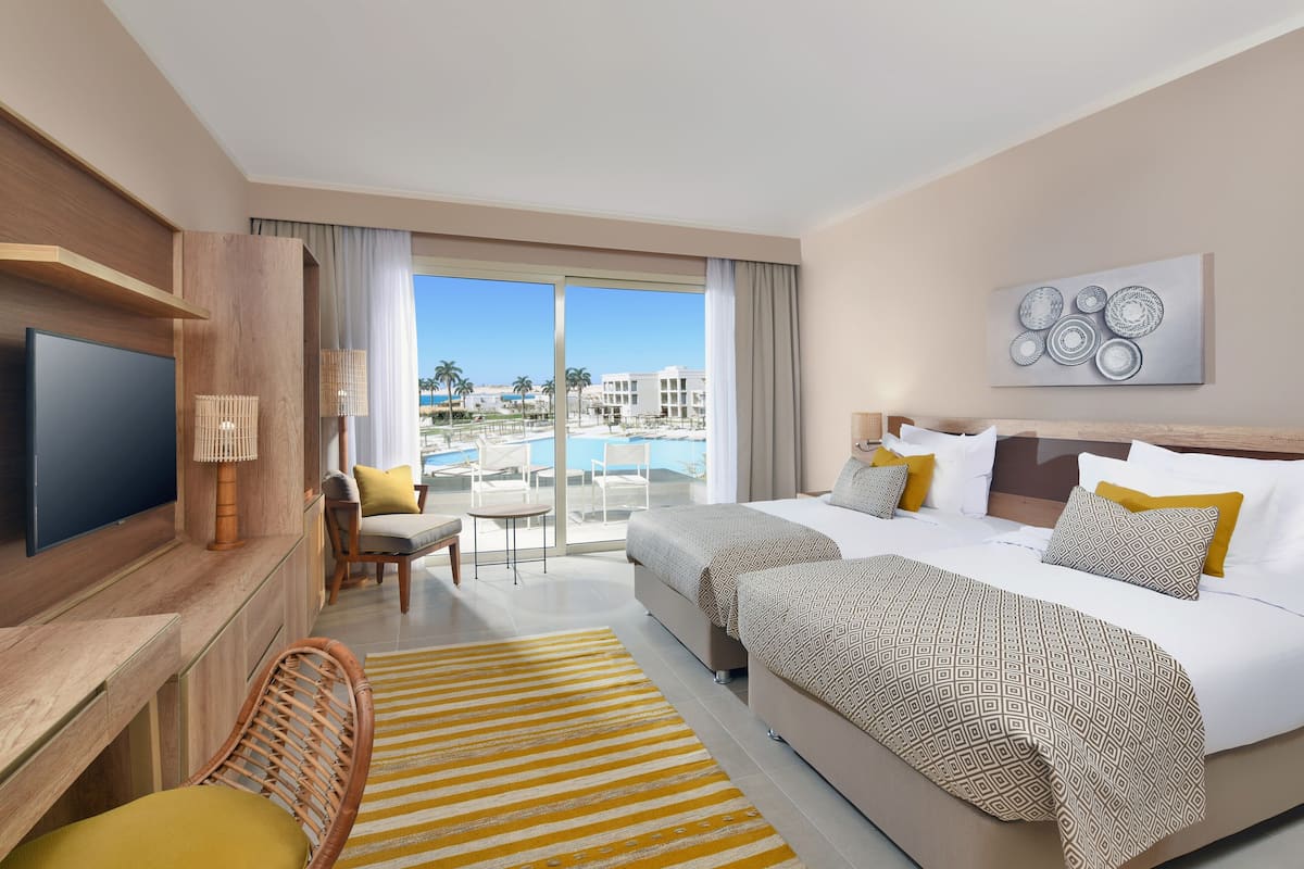 Quarto de hotel com uma cama grande, televisão, escrivaninha e vista para uma piscina e edifícios.