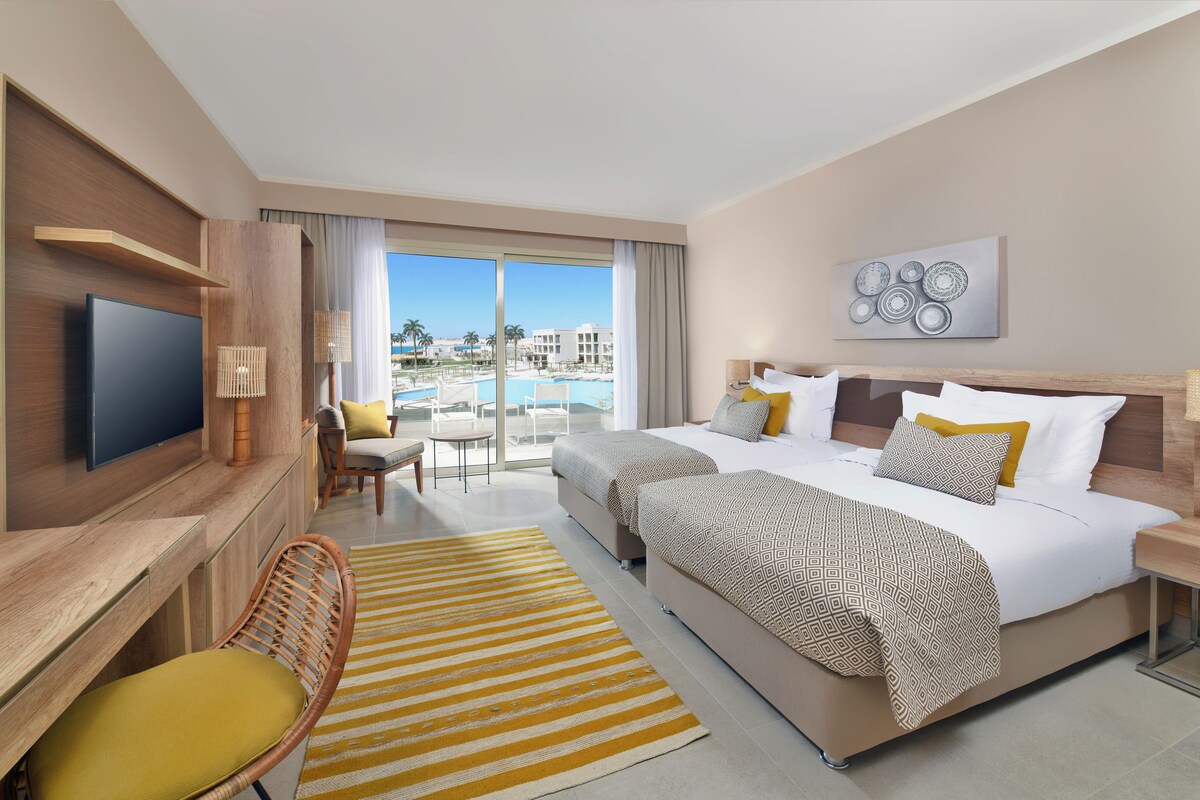 A hotel room with a large bed, a TV, a desk, a chair, and a view of a pool and buildings.
