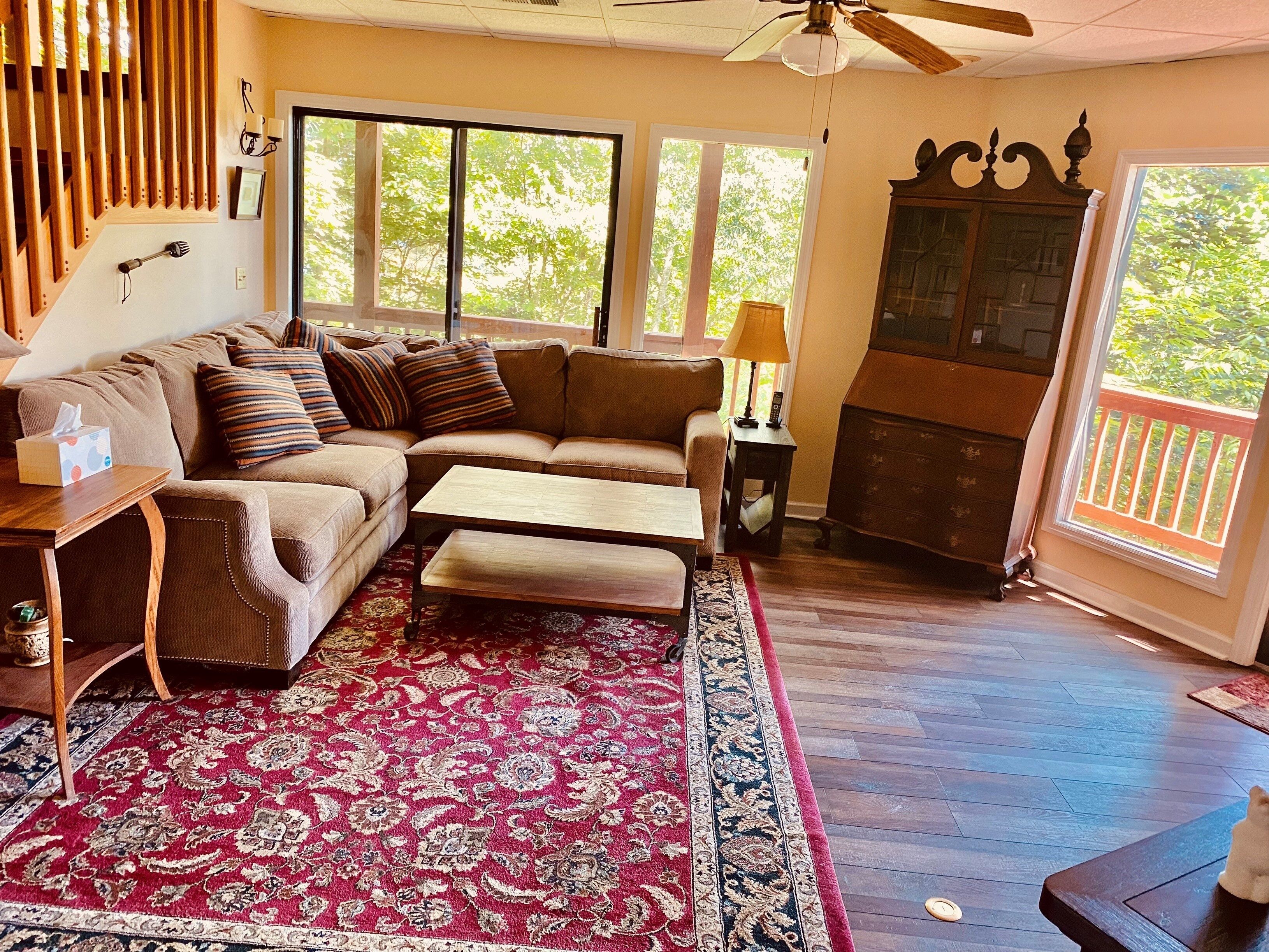A cozy living room with a sectional sofa, a wooden coffee table, a patterned rug, and a view of the outdoors through large windows.