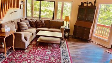 A cozy living room with a sectional sofa, a wooden coffee table, a patterned rug, and a view of the outdoors through large windows.