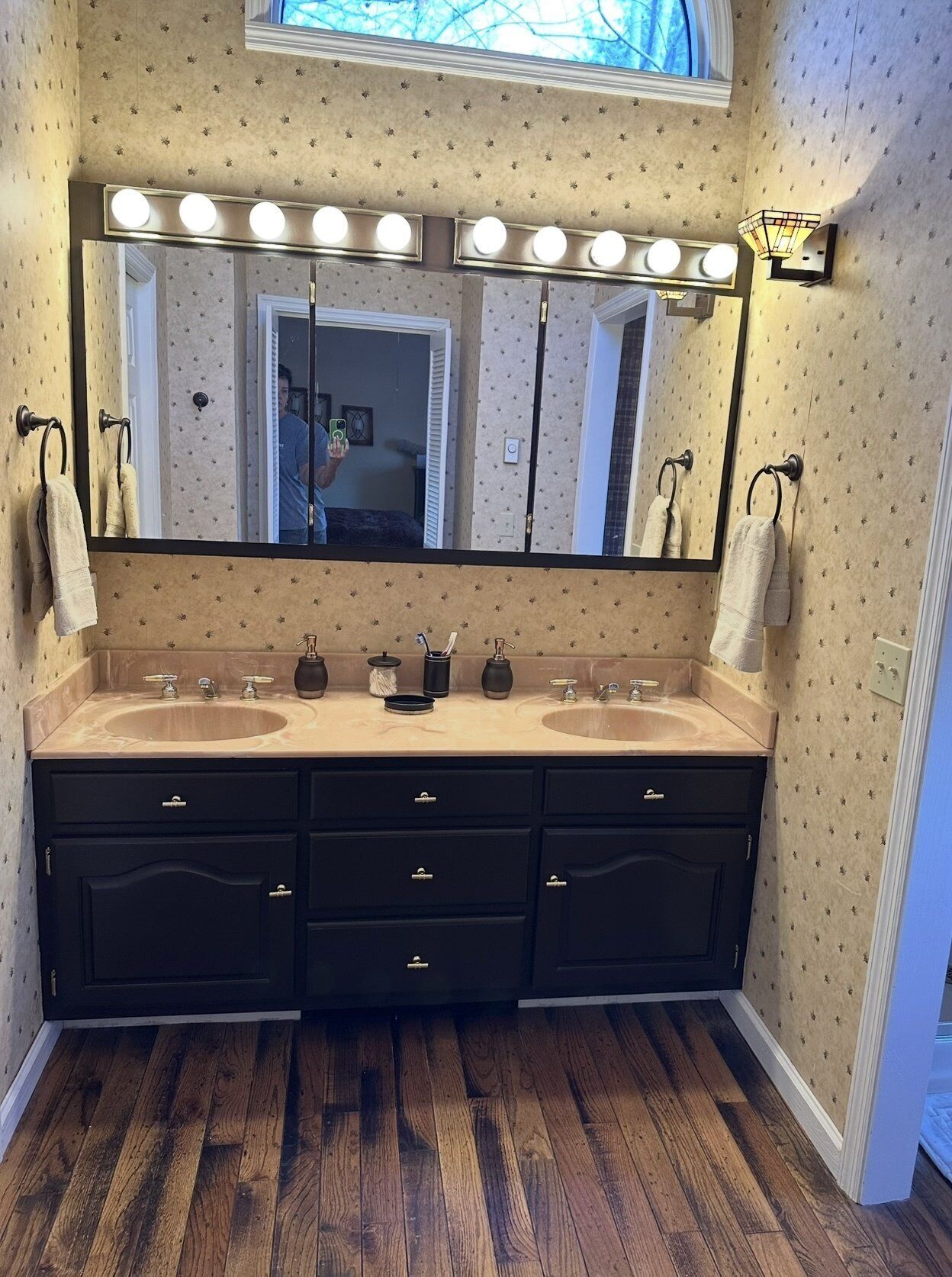 A bathroom with two sinks, a vanity with drawers, a large mirror with lights, and patterned wallpaper.