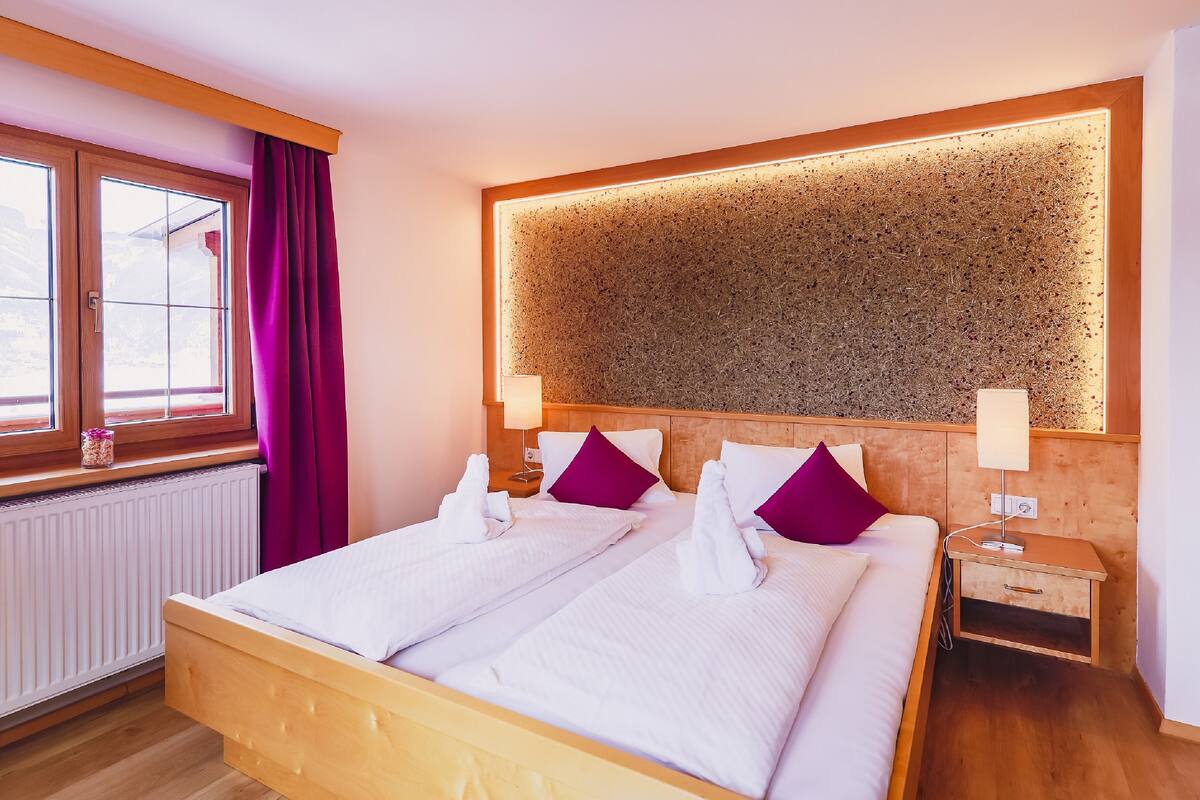 Quarto de hotel com cama de madeira, duas almofadas roxas, mesa de cabeceira com abajur e parede texturizada grande acima da cama.