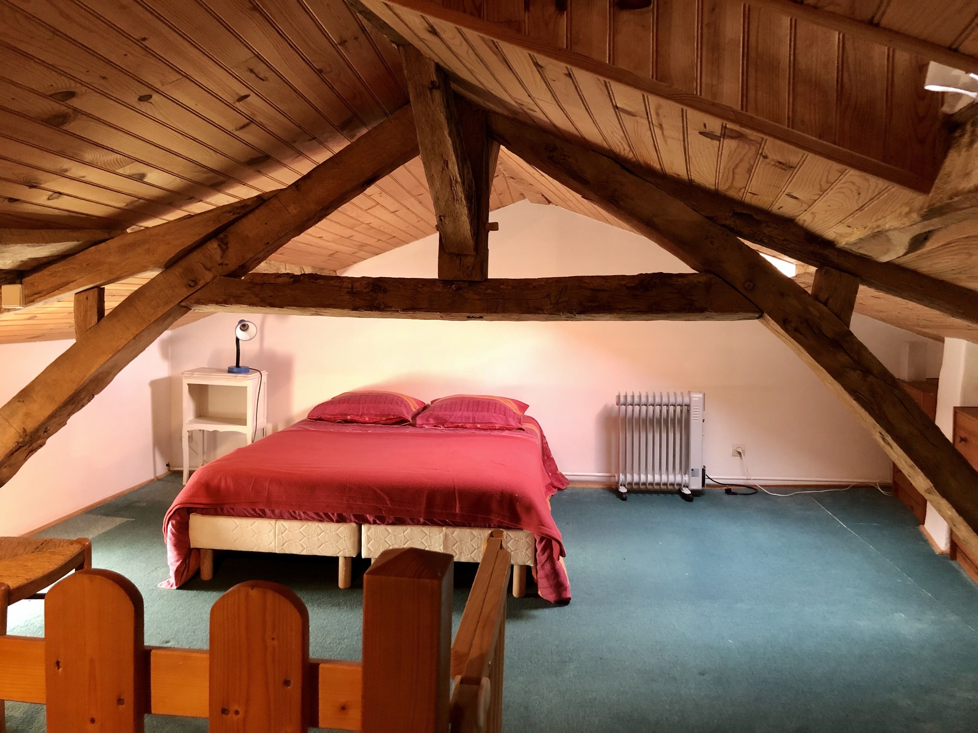 A bedroom with a wooden ceiling, a bed with red bedding, a white bedside table, and a radiator.