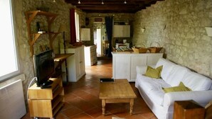Un salon de style rustique avec un canapé blanc, une table basse en bois et un téléviseur posé sur un meuble en bois. On aperçoit à l’arrière-plan un coin cuisine avec des murs en pierre et des poutres apparentes.