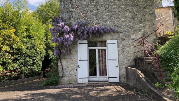 Un bâtiment en pierre doté d’une porte et de fenêtres, entouré de verdure et agrémenté d’un escalier.