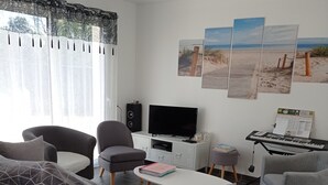 A modern living room with a grey sofa, two armchairs, a coffee table, a TV, a keyboard, and a wall with three beach-themed paintings. - 3* HOUSE WITH GARDEN AND TERRACE (sleeps 8) in Pont-L'Abbe (Pont-l'Abbé)