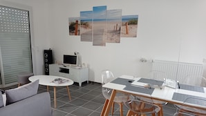 A modern living room with a grey sofa, a white coffee table, and a dining area with a white table and clear chairs. A large beach-themed artwork is displayed on the wall. - 3* HOUSE WITH GARDEN AND TERRACE (sleeps 8) in Pont-L'Abbe (Pont-l'Abbé)