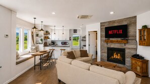 A modern living room with a fireplace, a large flat-screen TV, and a dining area with natural light.