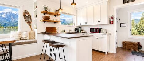 Una cocina con armarios blancos, una isla de madera, taburetes de bar, una lámpara colgante y vistas a las montañas.