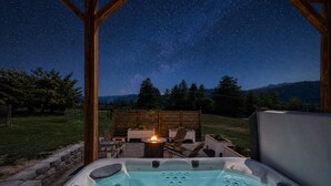 A hot tub with a fire feature, surrounded by wooden deck furniture, under a starry night sky.