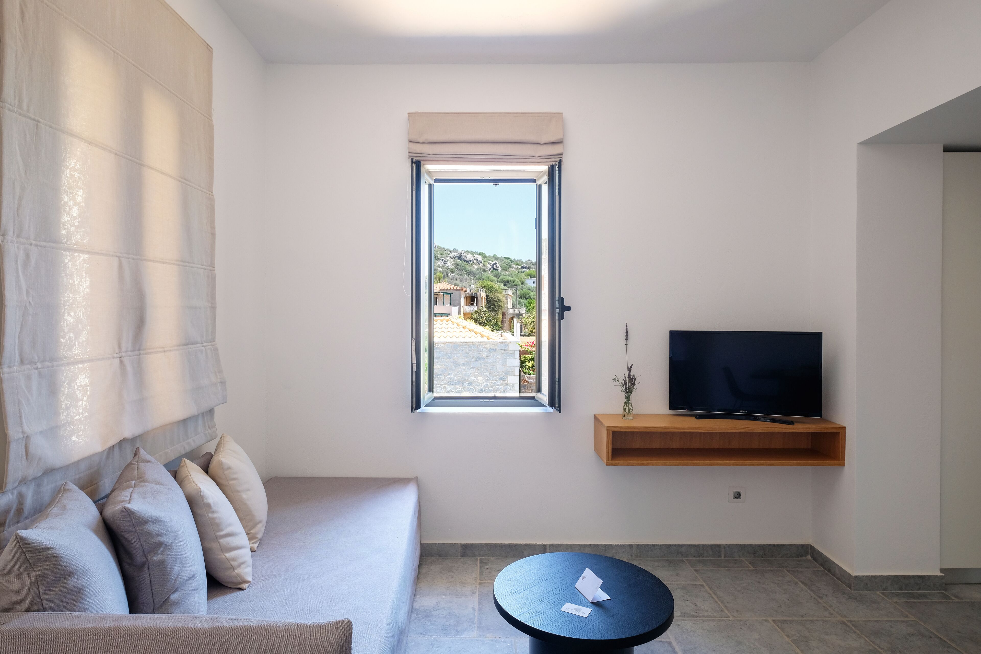 Una sala de estar moderna con un sofá, una pequeña mesa redonda, un televisor sobre una estantería de madera y una ventana con vistas a edificios y colinas.