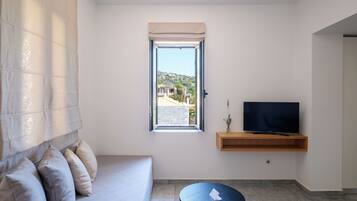 A modern living room with a sofa, a small round table, a television on a wooden shelf, and a window with a view of buildings and hills.