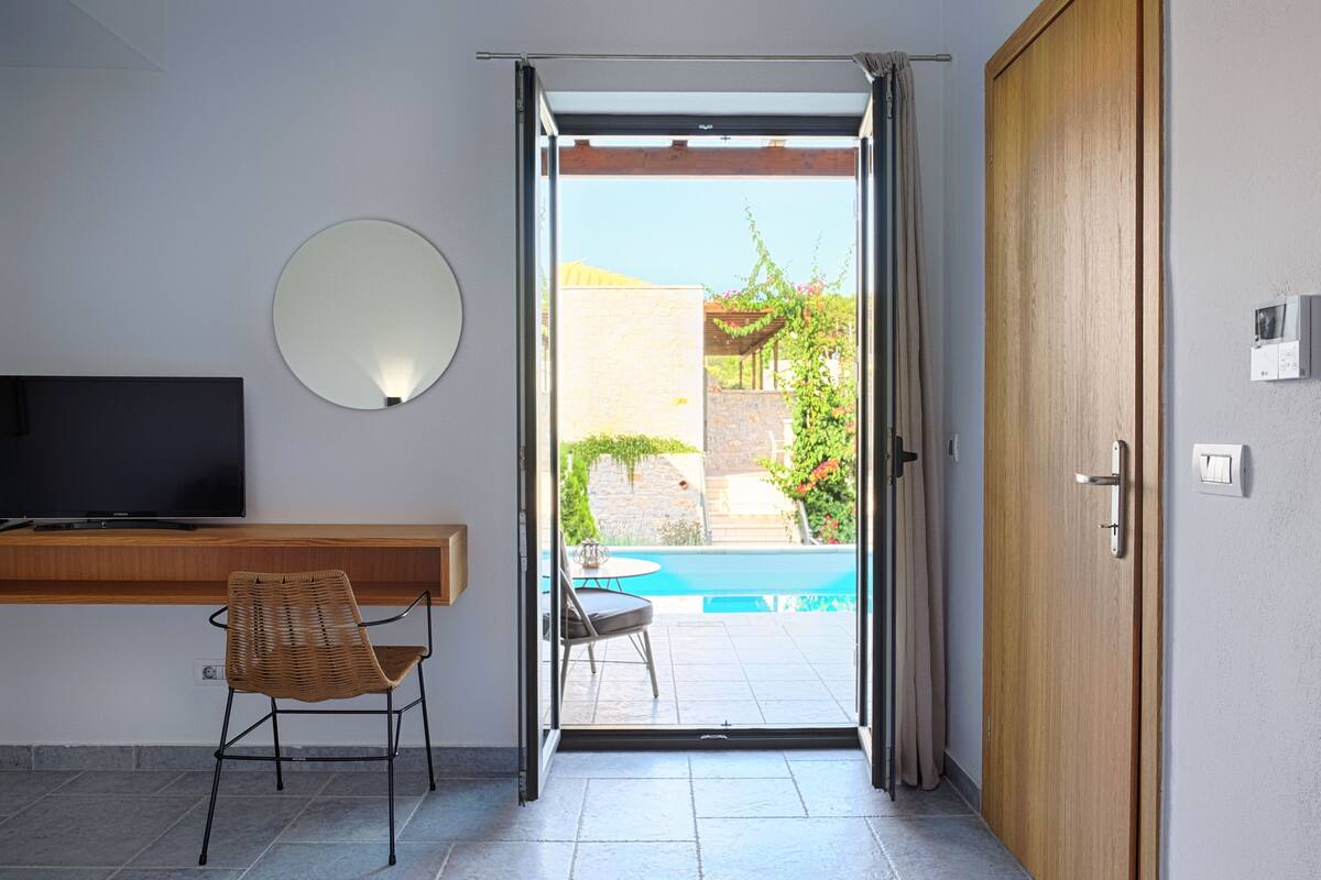 A room with a TV, a wicker chair, a door leading to a pool area, and a wooden door.