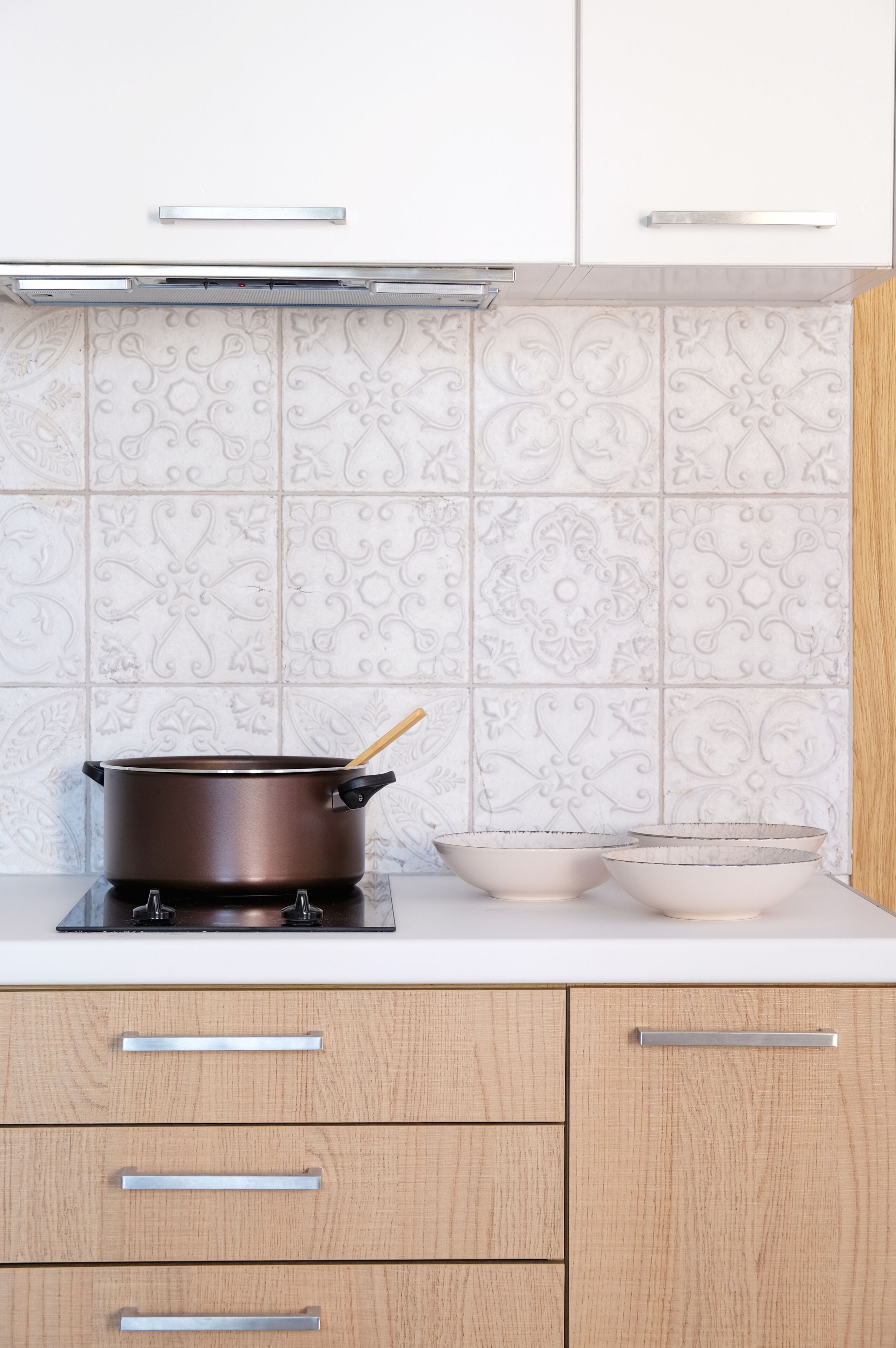 A kitchen with a stove, pots, and bowls on the counter, light wooden cabinets, and decorative tiles on the backsplash.