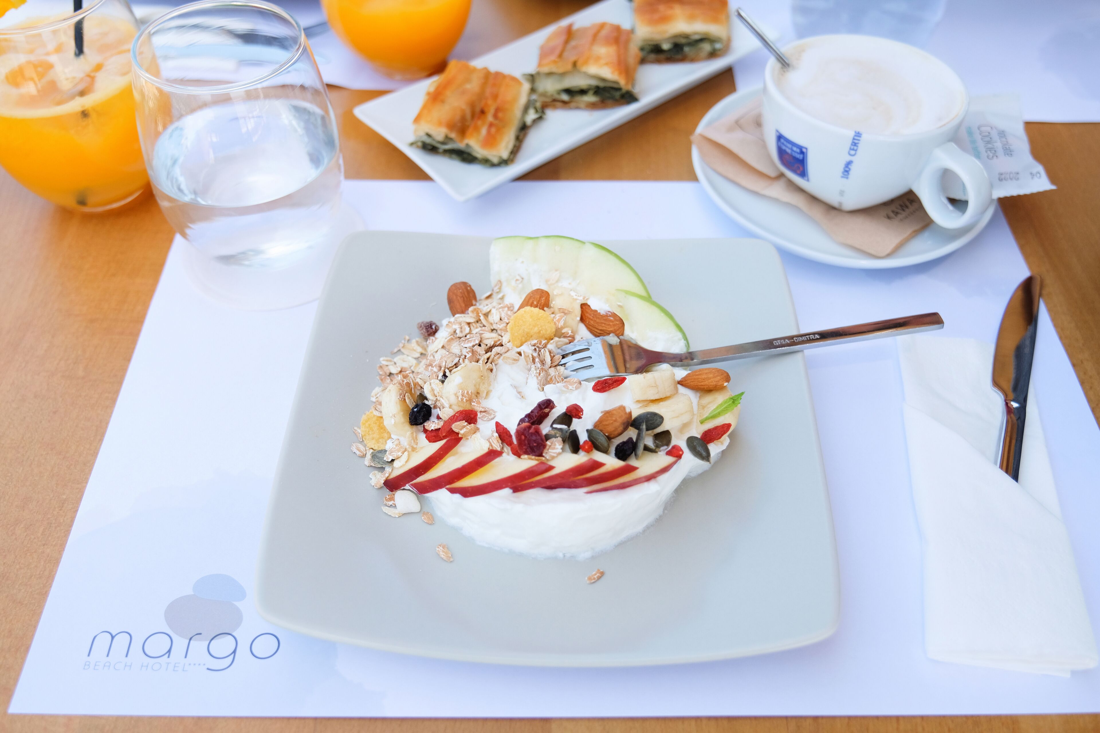 Un desayuno con un plato de yogur cubierto de frutas y nueces, un vaso de agua, una taza de café y una porción de bollería sobre una mesa.