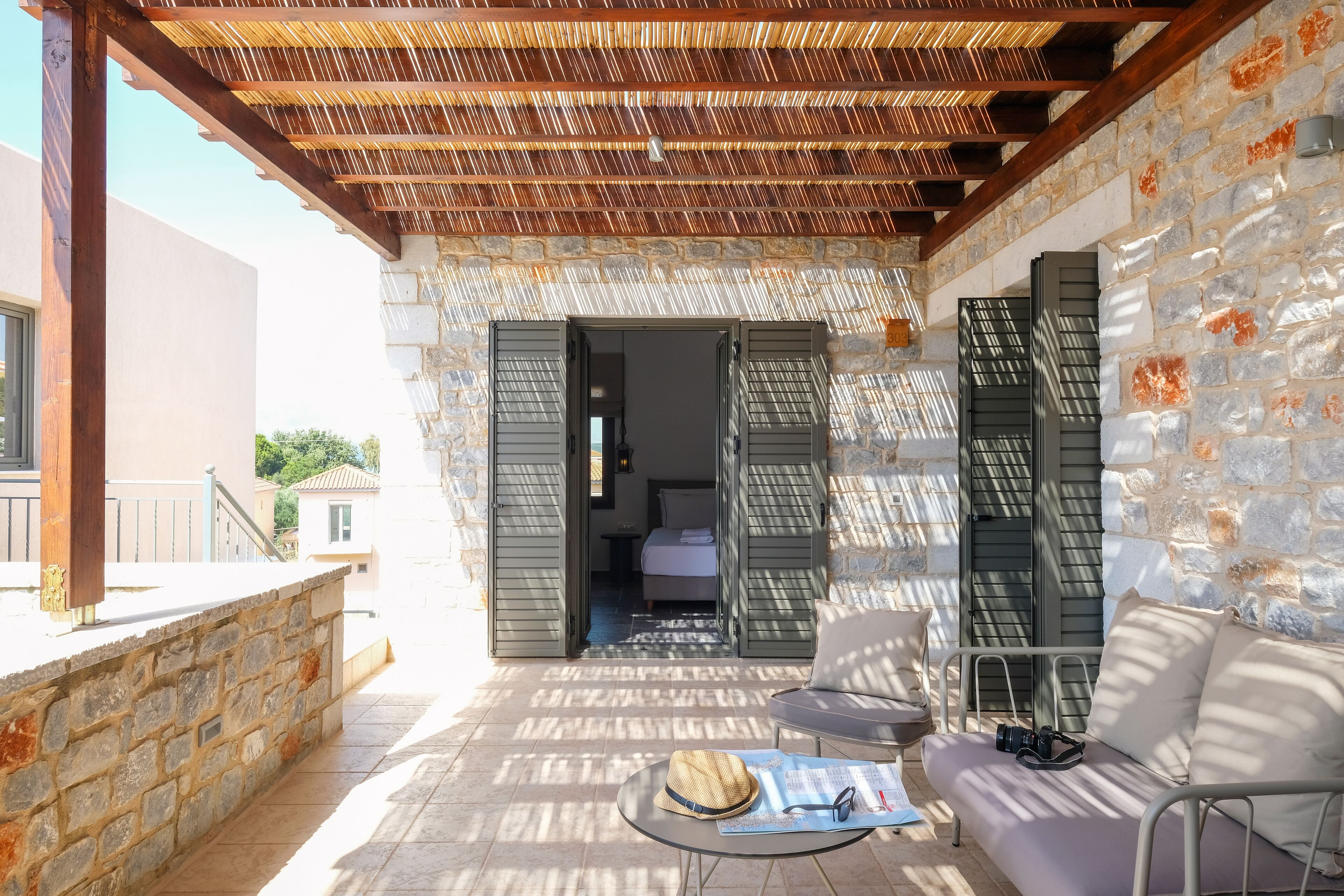 A sunlit patio with stone walls, a wooden pergola, and a sofa with a hat and sunglasses on a table.
