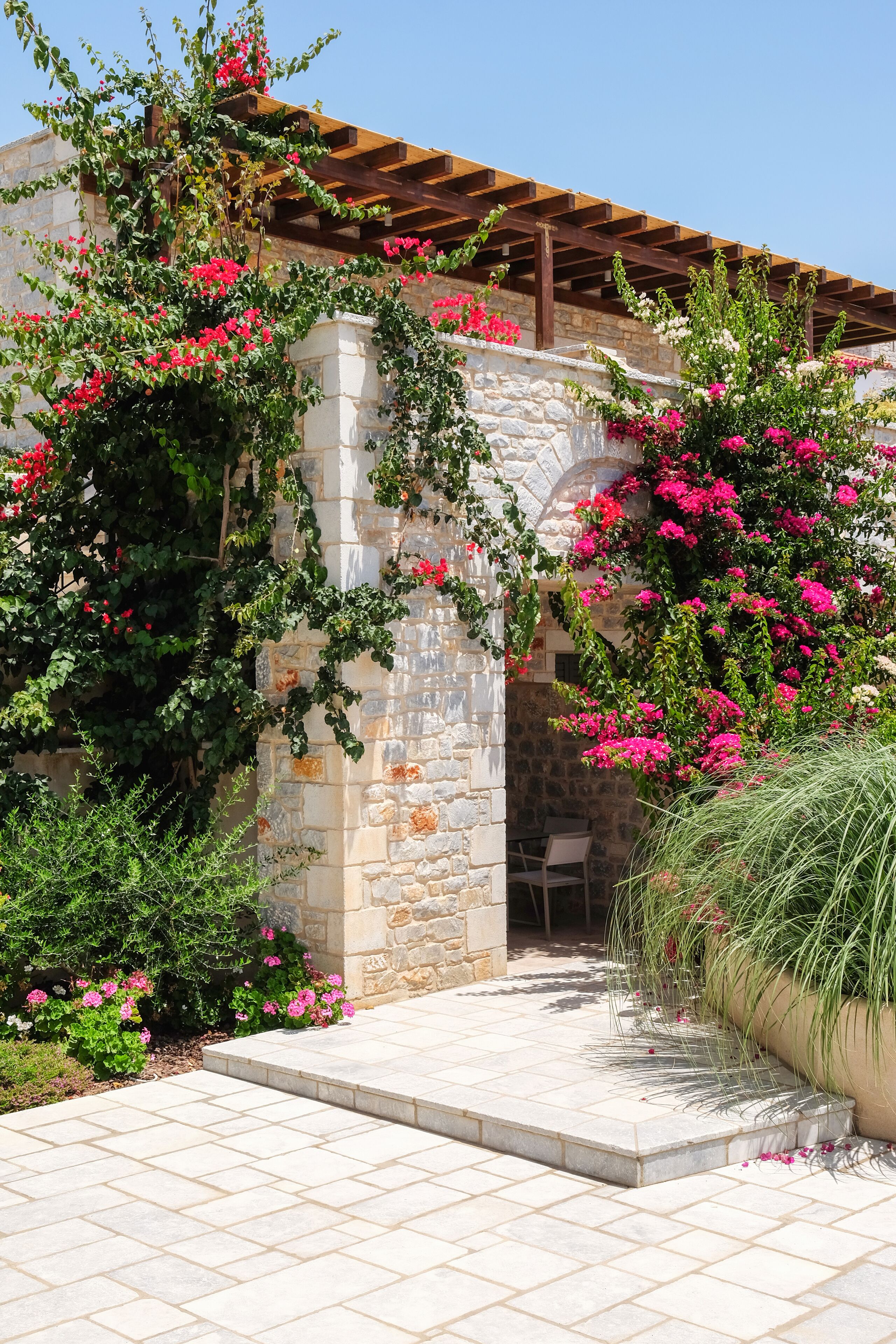 A stone archway with climbing plants and flowers, leading to a covered outdoor seating area.