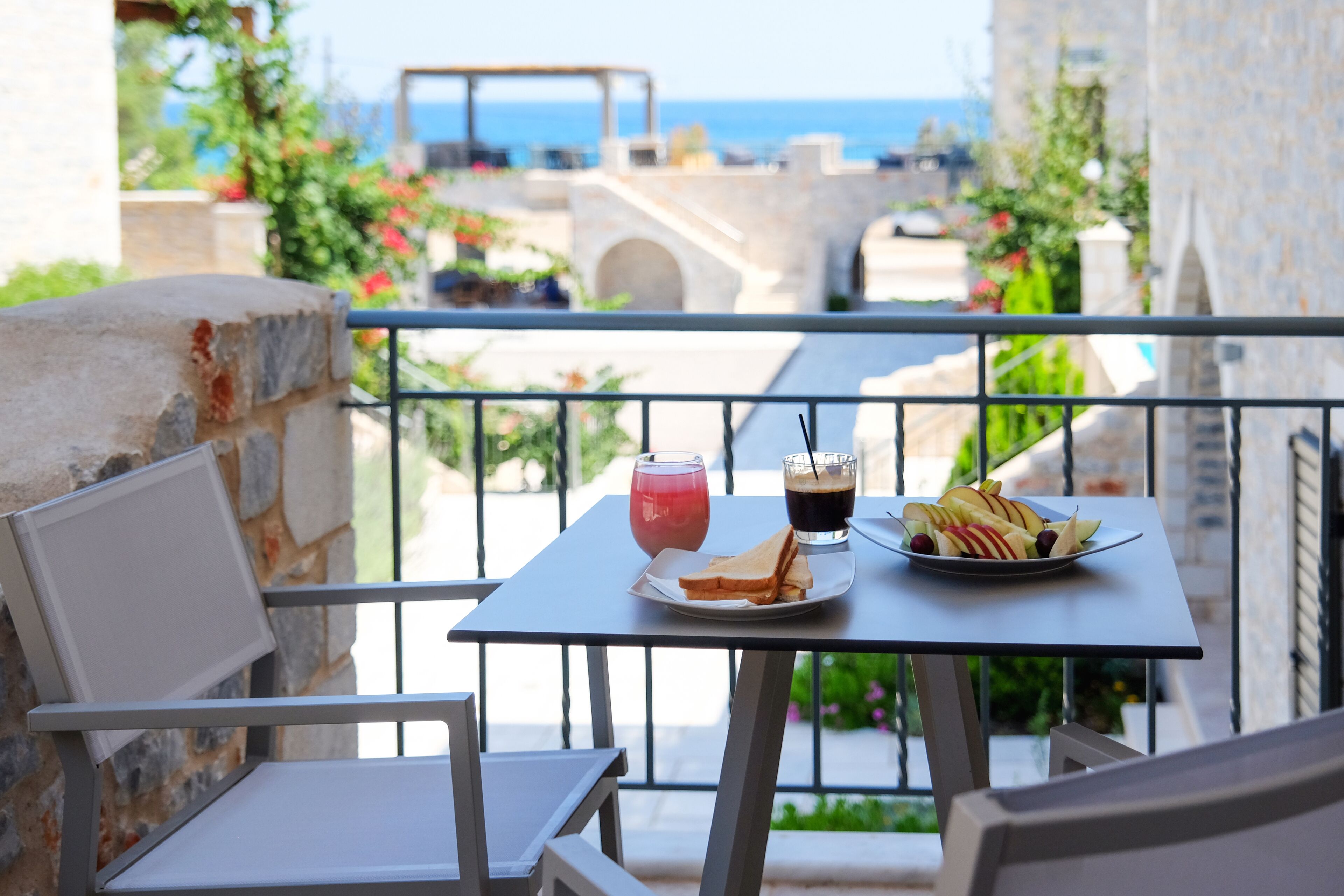 Un balcón con una mesa dispuesta para comer, que incluye un plato de fruta y dos bebidas, con vistas panorámicas al mar y edificios.
