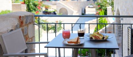 Un balcón con una mesa dispuesta para comer, que incluye un plato de fruta y dos bebidas, con vistas panorámicas al mar y edificios.