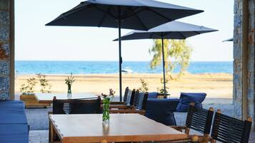 Un patio frente a la playa con una mesa y sillas de madera, dos sombrillas y vistas al océano.