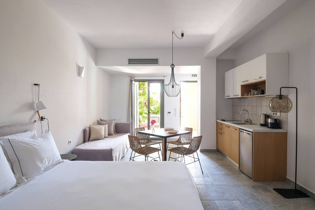 A modern hotel room with a large bed, a dining table with chairs, a kitchenette, and a view of the outdoors.