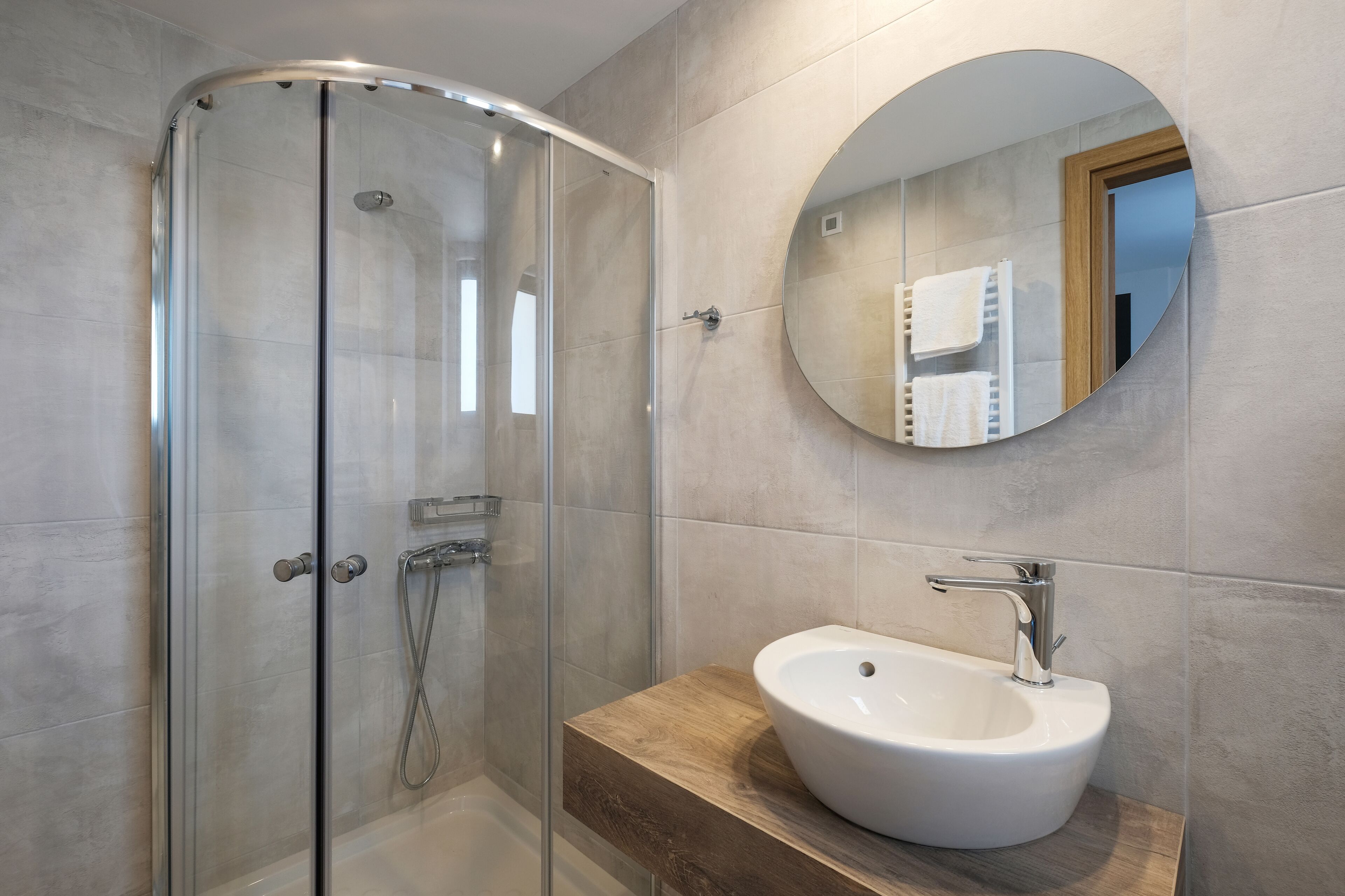 A modern bathroom with a glass shower enclosure, a round mirror, and a white sink with a wooden countertop.