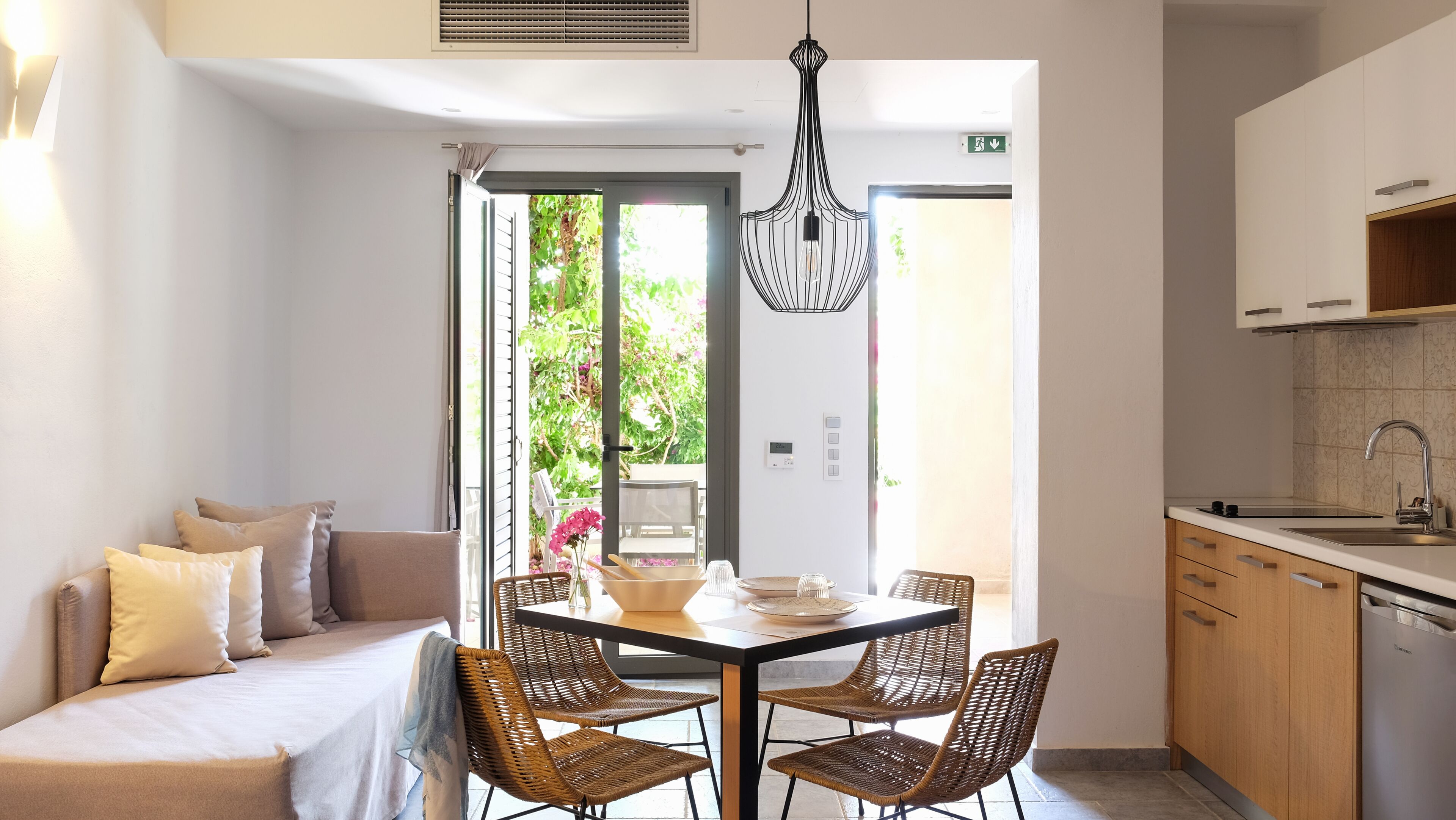 A modern room with a bed, a dining table with chairs, a kitchen area, and a view of greenery outside.