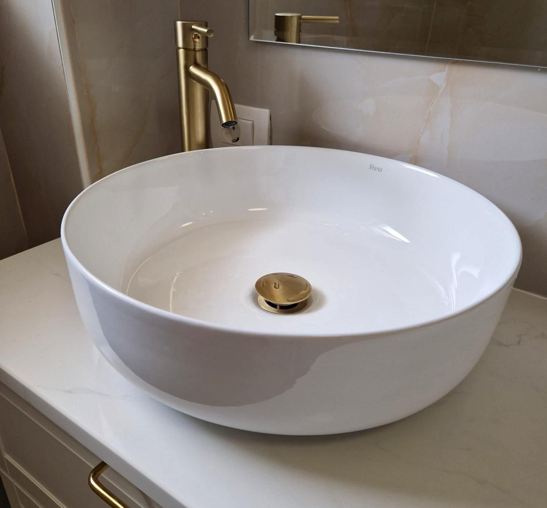 A white vessel sink with a gold faucet on a white countertop.