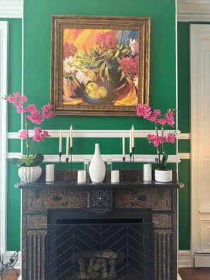 A fireplace with a floral painting above it, a white vase, and pink orchids on the mantel.