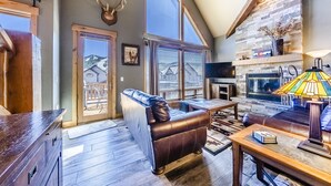 A cozy living room with a leather sofa, a fireplace, and a large window overlooking a snowy landscape. - Spacious Townhome with Private Hot Tub, minutes away from Winter Park Resort (Winter Park)