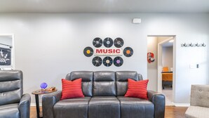 Un salon avec un canapé en cuir, des coussins rouges, une table basse ornée d’une plante, et un mur décoré de disques vinyle et d’un panneau indiquant « MUSIQUE ».