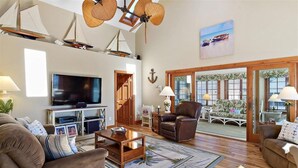 A spacious living room with a large flat-screen TV, leather armchairs, a wooden coffee table, and a view of the outdoors through a sliding glass door.