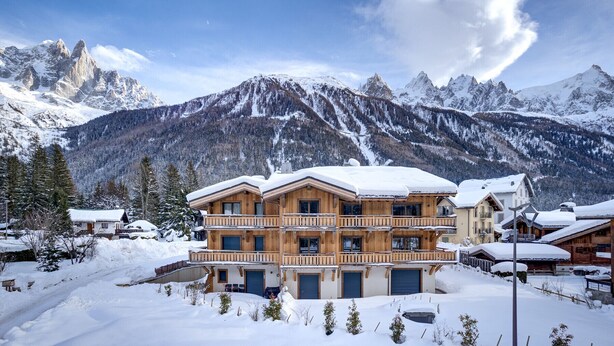 Een meerlaags houten chalet met balkons, omgeven door besneeuwde bergen en bomen.
