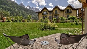 Een hotel met een houten terras en twee stoelen, met uitzicht op een weelderig groen gazon en een bergketen.