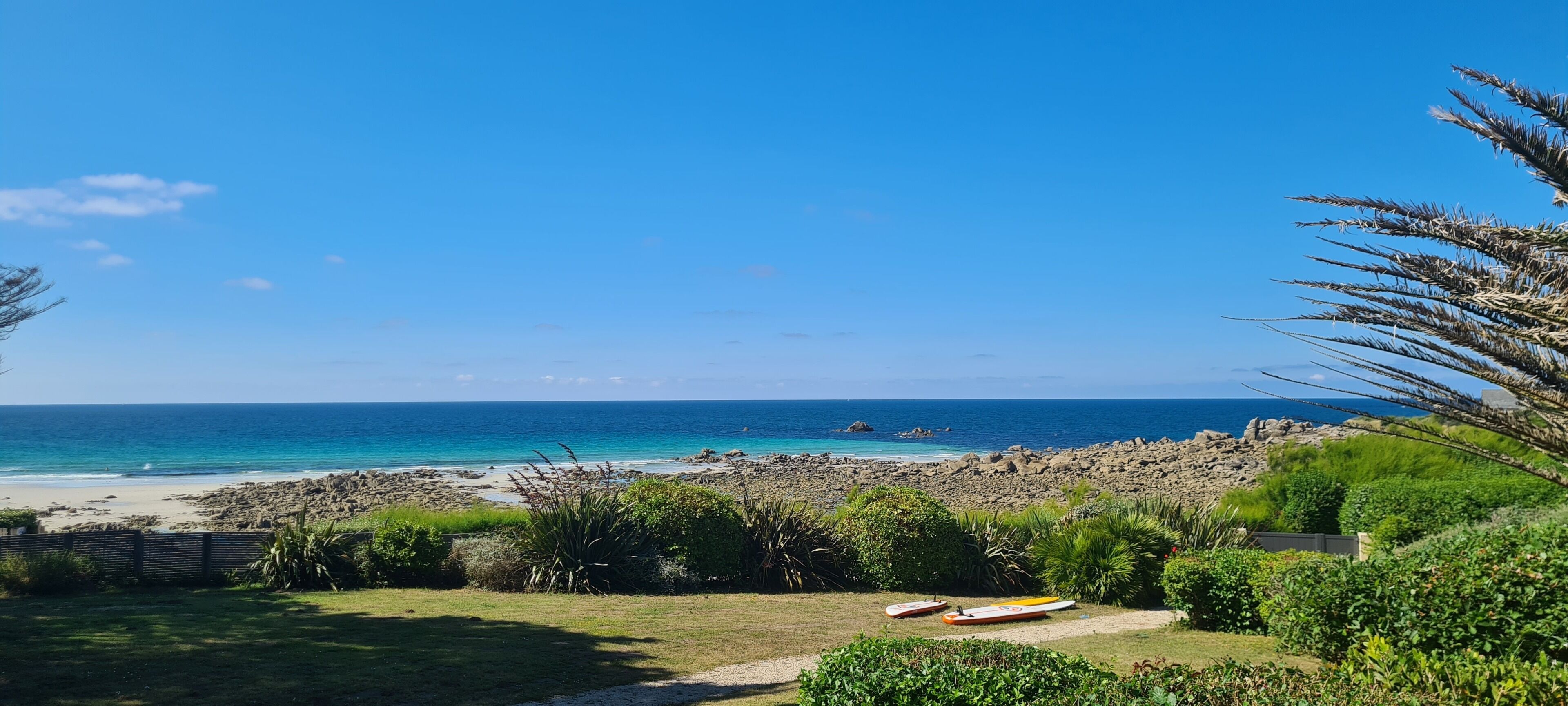 Une plage avec une eau d’un bleu limpide, un rivage rocheux et une zone herbeuse où se trouve une planche de surf.