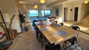 Un vaste espace dédié à la salle à manger, avec une table et des chaises en bois, un grand vase décoratif servant de centre de table, et une vue sur l’océan grâce à de grandes fenêtres.