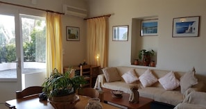 Una sala de estar con sofá, una mesa de centro, una mesa de comedor y sillas. Hay plantas en macetas y cuadros en las paredes.
