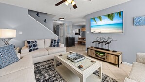 A living room with a ceiling fan, a TV displaying a beach scene, a sofa with patterned cushions, and a coffee table with decorative items.