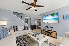 Uma sala de estar com um ventilador de teto, uma TV exibindo uma cena de praia, um sofá com almofadas estampadas e uma mesa de centro com objetos decorativos.