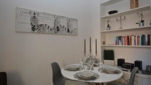 Een eetruimte met een ronde tafel gedekt voor vier personen, omringd door grijze stoelen. Een kandelaar met drie kaarsen staat op tafel. Daarboven hangt een groot zwart-wit abstract kunstwerk. Aan de rechterkant staat een boekenkast met verschillende boeken en decoratieve objecten.
