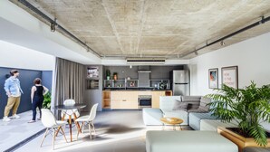 Ein modernes Wohnzimmer mit einer Couch, Esstisch und Stühlen, einer Küchenzeile mit Schränken und Geräten sowie einem großen Fenster, das viel natürliches Licht hereinlässt.